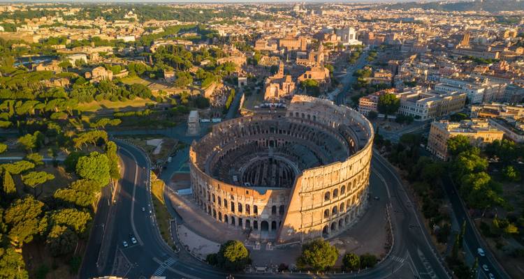 Dramatische Luftaufnahme des Kolosseums, umgeben von der weitläufigen Stadtlandschaft Roms im warmen Sonnenlicht des Sonnenaufgangs.