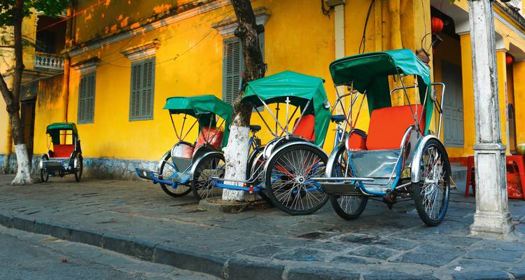 Bunte Rikschas, die vor einem gelben Gebäude in einer vietnamesischen Umgebung geparkt sind.