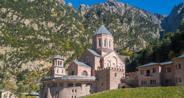 Stone monastery complex set against steep forested mountains under clear blue sky.