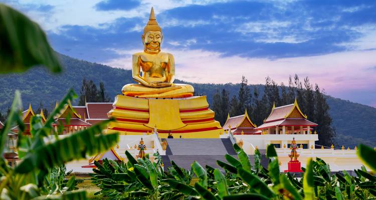 Grande statue dorée de Bouddha assis domine un complexe de temple au sommet d'une colline avec des toits thaïlandais traditionnels contre un ciel coloré du soir et des collines boisées.