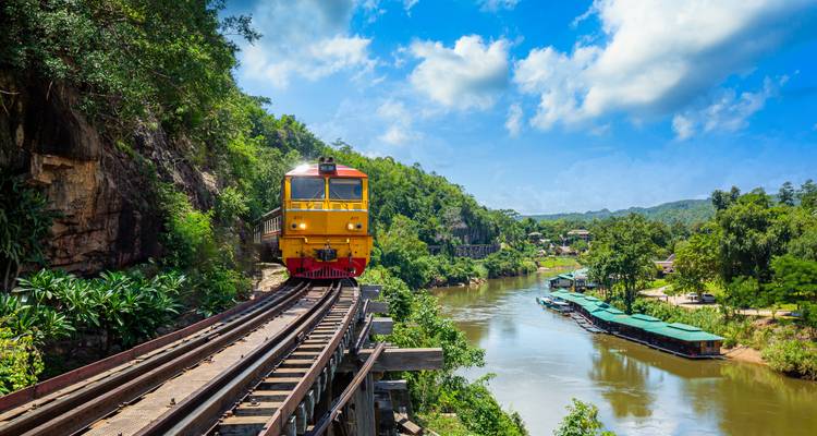 Un train jaune vif traverse un pont de chevalets en bois haut au-dessus d'une rivière sinueuse et d'une jungle luxuriante sous un ciel bleu éclatant.