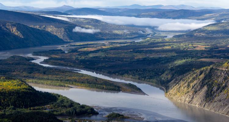 Weites Flusstal mit gewundenen Wasserwegen, bewaldeten Hängen und fernen nebligen Bergen.