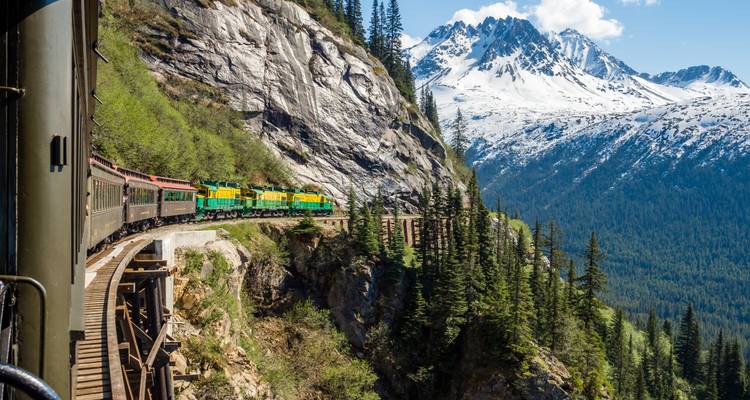 Historische Schmalspurbahn, die sich entlang einer Klippe windet, mit schneebedeckten Gipfeln, die dahinter aufragen.