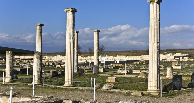 Überreste alter Steinsäulen und Fundamente, die auf einer offenen archäologischen Stätte unter klarem Himmel stehen