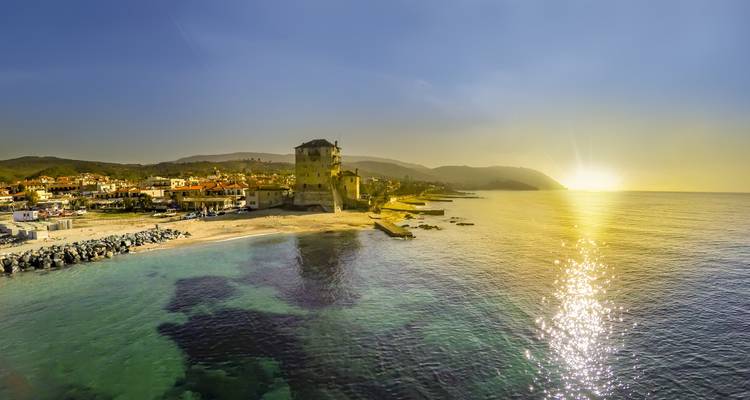 Luftaufnahme einer Küstenlandschaft mit einem Küstendorf und mittelalterlichem Turm bei Sonnenuntergang mit goldenen Reflexionen auf dem Wasser