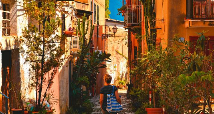 Person, die in einer schmalen Gasse einer Altstadt mit üppigem Grün spaziert.