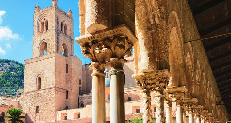 Nahaufnahme von kunstvoll verzierten Kreuzgangssäulen mit dem Glockenturm der Kathedrale von Monreale im Hintergrund.
