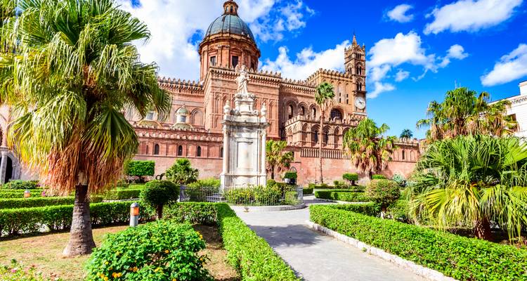 Palmengesäumter Garten, der zur kuppelgekrönten und zinnenbesetzten Fassade der Kathedrale von Palermo unter blauem Himmel führt.