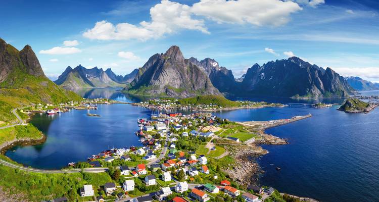 Panoramablick aus der Luft auf ein malerisches Fjorfdorf, umgeben von steilen Bergspitzen und glitzerndem blauem Wasser unter einem klaren Himmel.