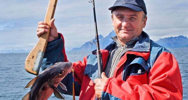 Ein älterer Mann in farbenfroher Outdoor-Kleidung präsentiert stolz einen frisch gefangenen Fisch auf einem Boot, während dramatische gezackte Berge hinter einem ruhigen Meer aufragen.