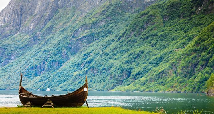 Ein altes hölzernes Wikinger-Boot ruht auf grünem Gras neben einem türkisfarbenen Fjord, der von steilen, mit Bäumen bewachsenen Klippen eingefasst ist.