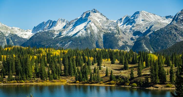 Des sommets montagneux saupoudrés de neige dominant une forêt de pins et un lac tranquille par temps clair.
