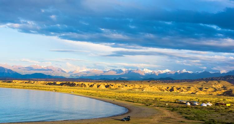 Plage incurvée sur un lac alpin tranquille avec des yourtes au loin et une chaîne de montagnes enneigées.