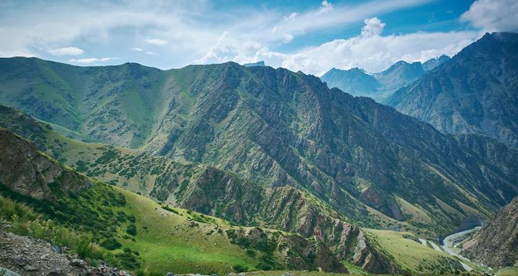 Des crêtes montagneuses vertes et rocheuses escarpées sous un ciel bleu partiellement nuageux en Asie centrale.
