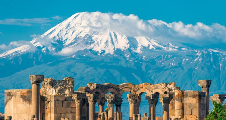 Ruinen der Zvartnots-Kathedralensäulen mit schneebedecktem Berg Ararat im Hintergrund unter blauem Himmel.