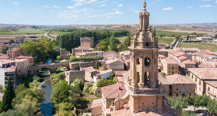 Vista aérea de un pueblo español medieval con una prominente torre de campanario de iglesia y viñedos y río circundantes.