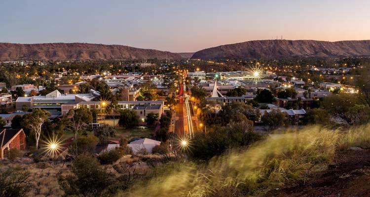 Cityscape at dusk with glittering lights and hills in the background.
