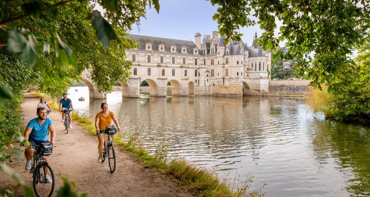 Los ciclistas recorren un camino junto al río enmarcado por árboles con el Château de Chenonceau de fondo.