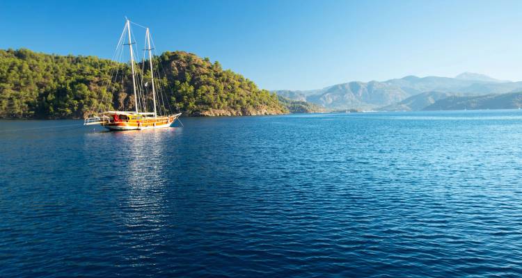 Velero gulet tradicional anclado en una tranquila bahía azul rodeada de colinas cubiertas de pinos verdes.