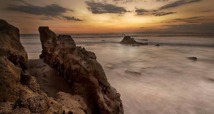Fotografía de larga exposición del paisaje marino con rocas escarpadas y olas neblinosas bajo un cielo cálido al atardecer.