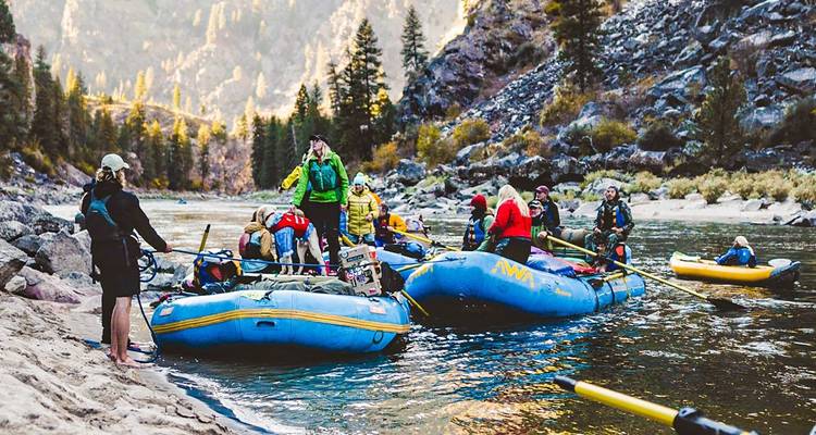 Los aventureros preparan balsas inflables azules en la orilla boscosa de un río rodeada por imponentes paredes de cañón.