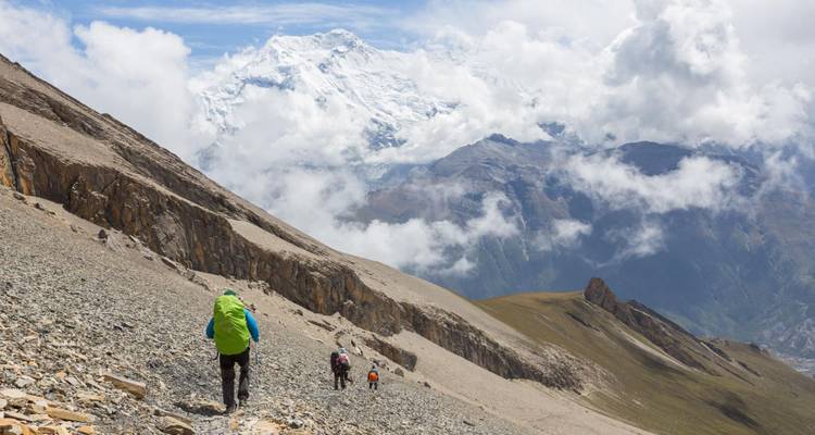 Groupe de randonneurs montant une pente rocheuse de montagne avec des pics enneigés spectaculaires et des nuages qui s'étirent devant eux.