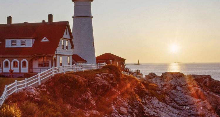 Faro histórico y casa del farero encaramados en la costa rocosa de Maine bañados por la cálida luz del atardecer con suaves olas del océano abajo.