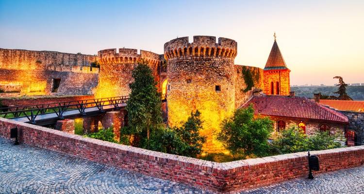 Forteresse historique avec tours de pierre et toits rouges au coucher du soleil.