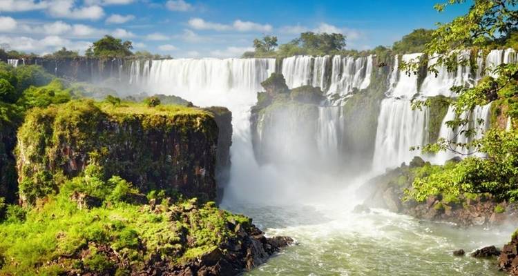 Foto panorámica iluminada por el sol de las imponentes Cataratas del Iguazú rodeadas de exuberante vegetación.
