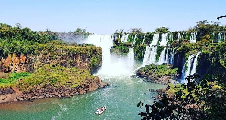 Vista amplia de múltiples cascadas de las Cataratas del Iguazú cayendo hacia el río con un barco de tour abajo.