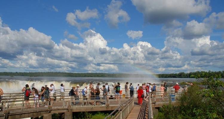 Las multitudes se reúnen en un paseo peatonal con vista a las Cataratas del Iguazú con niebla y un débil arco iris bajo un cielo nublado.
