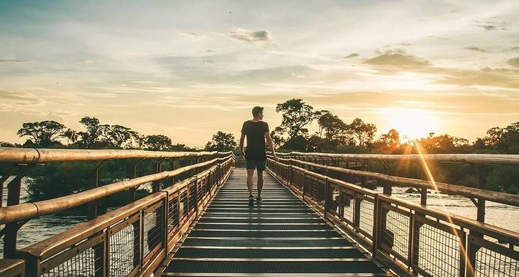 Un viajero solitario camina por un puente metálico al atardecer con la luz dorada reflejándose en el río Iguazú abajo.