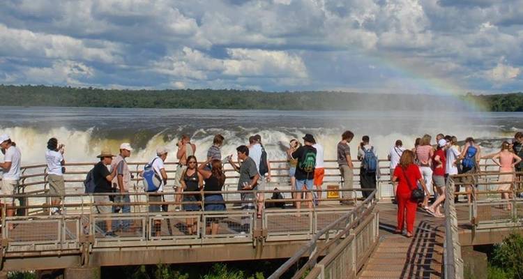 Los visitantes se paran en una plataforma junto a las atronadoras Cataratas del Iguazú mientras un arcoíris se arquea a través de la bruma.