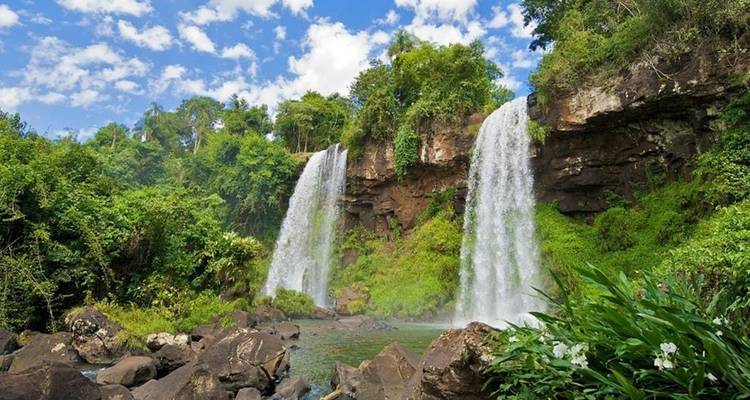 Dos cascadas caen en picada hacia una piscina de la selva bajo un cielo azul brillante con nubes dispersas.
