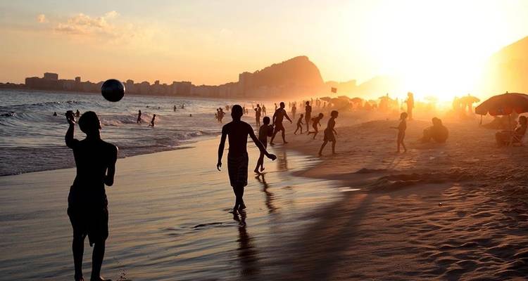 Am Strand von Rio silhouettierte Strandbesucher, die bei goldenem Sonnenuntergang Fußball spielen und schwimmen
