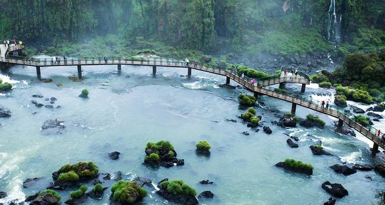 Luftaufnahme einer geschwungenen Brücke mit Touristen über wirbelnden türkisfarbenen Gewässern in der Nähe von Wasserfällen.
