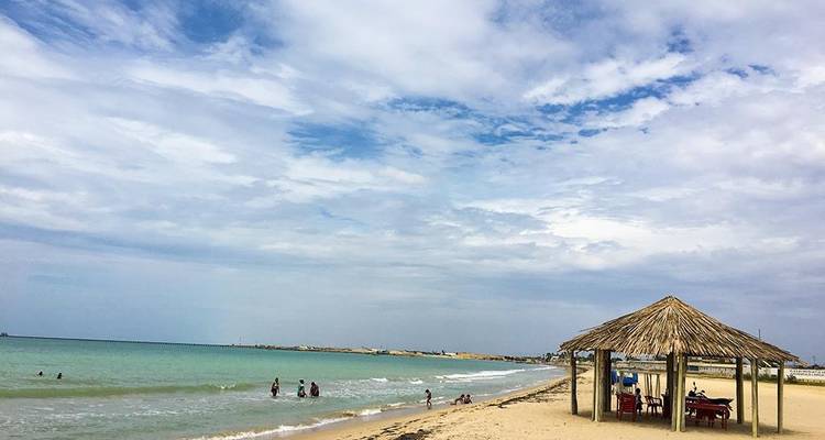 Een rustig Caribisch strand met een rieten hutje, zachte golven en verspreide zwemmers onder een wijde hemel.