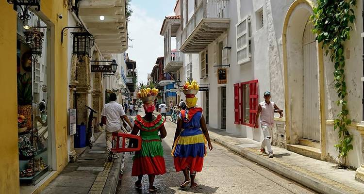 Kleurig geklede fruitverkopers en voetgangers lopen door een smal coloniaal straatje in de oude stad van Cartagena.