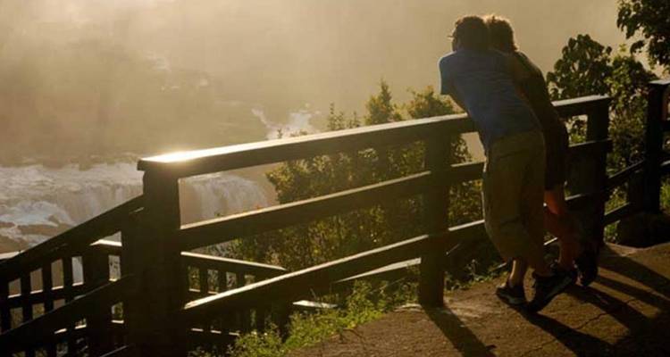 Een stel leunt tegen een houten reling en bewondert de Iguazu-watervallen in warm goudlicht.
