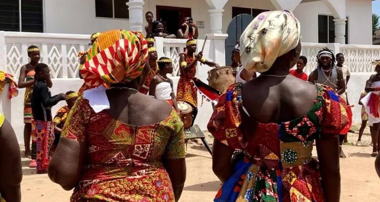 Des femmes vêtues de robes aux motifs éclatants regardent une performance dynamique de tambours et de danse villageoise devant un bâtiment blanc.
