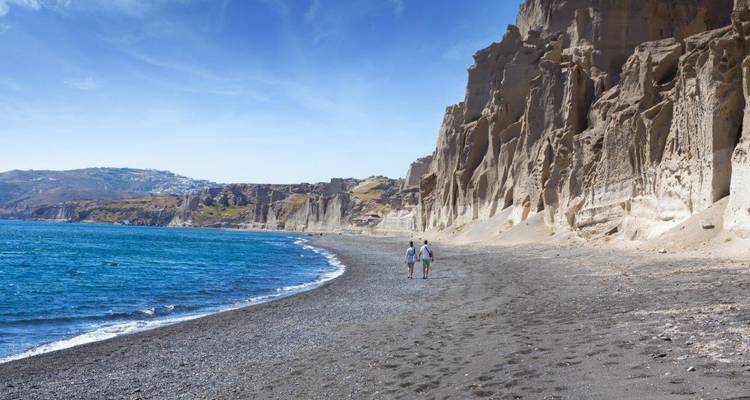 Pareja caminando en una playa con acantilados dramáticos y mar azul.