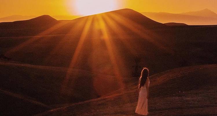 Une femme seule vêtue d'une robe blanche vaporeuse regarde vers le soleil levant projetant des rayons dorés sur les dunes du désert ondulantes.