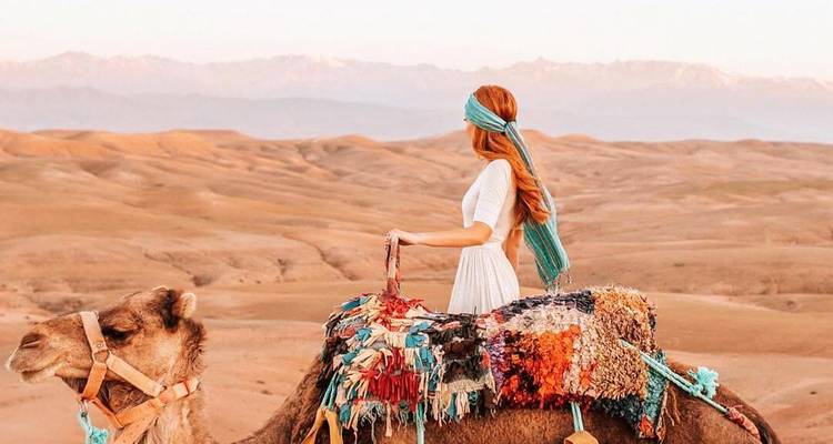 Femme aux cheveux roux vêtue d'une robe blanche chevauche un chameau orné à travers une plaine désertique ocre avec des montagnes enneigées au loin.