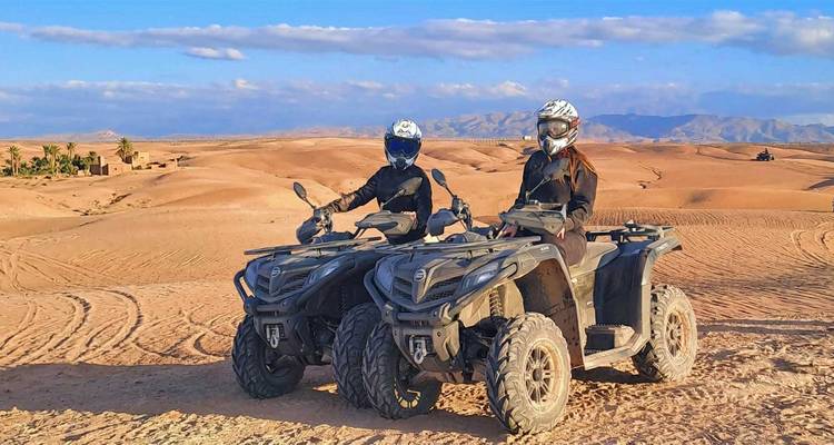 Deux voyageurs portant des casques posent sur des quads dans un vaste désert pierreux avec des montagnes au-delà