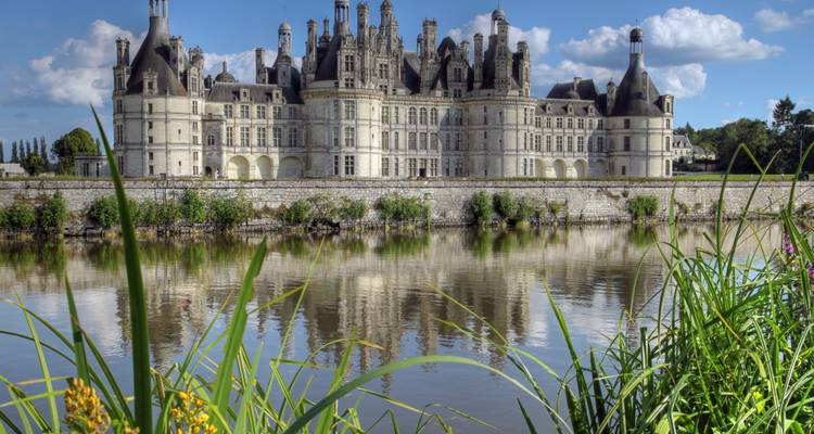 Château majestueux avec plusieurs tours et une architecture complexe se reflétant sur un plan d'eau serein, encadré de verdure.