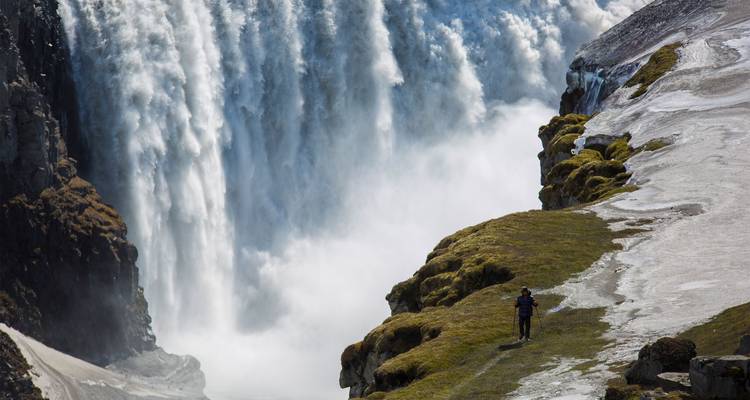 Einzelner Wanderer, winzig neben dem tosenden Vorhang eines gewaltigen Wasserfalls, der neben einer moosbedeckten Felswand herabstürzt