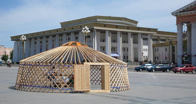Traditional yurt structure in front of a large building and cars.