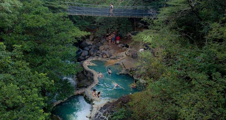 Personas disfrutando de una piscina natural en un bosque con un puente colgante arriba.