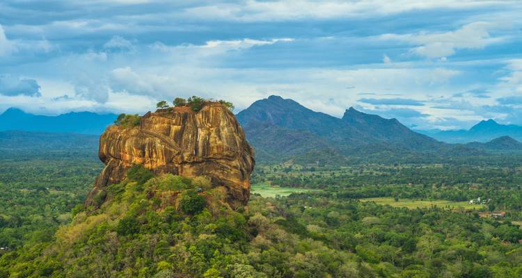 Majestic Sigiriya Rock Fortress towers over verdant forest and distant blue mountains under dramatic clouds.