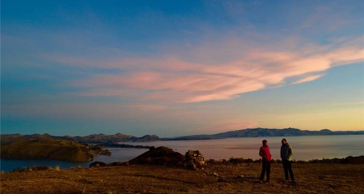 Zwei Reisende unterhalten sich auf einem Hügel bei Sonnenuntergang mit Blick auf die ausgedehnten Gewässer des Titicaca-Sees.